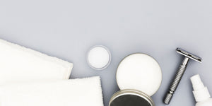 Flat lay of shaving essentials including post-shave balm, a safety razor, and white towels on a gray background, illustrating what post-shave balm is used for.