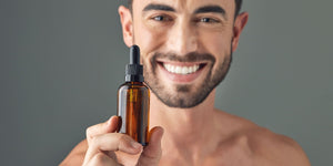 Close-up of a smiling man holding a bottle of beard oil, showcasing the product in focus. Ideal representation for a guide on when to start using beard oil, highlighting the benefits of proper beard care. 