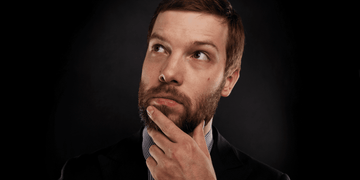 A bearded man in a suit is thoughtfully looking upward with his hand on his chin against a dark background. His expression suggests he is pondering the question, Why did my beard stop growing?