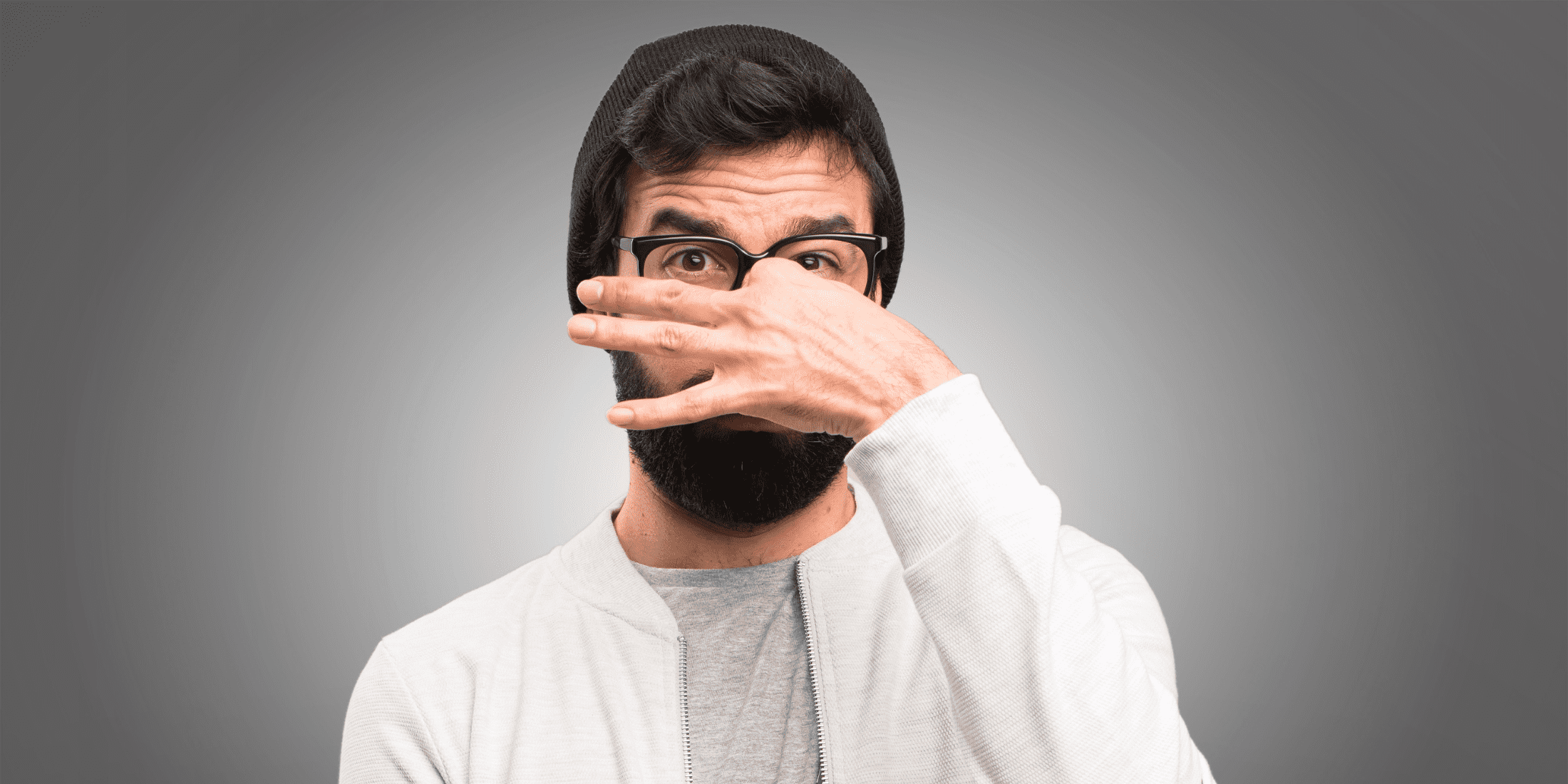 Young Man Wearing Glasses and a Beanie, Holding His Nose Due to Strong Beard Odor - Learn Expert Tips to Prevent and Eliminate Bad Beard Smells and Maintain Freshness