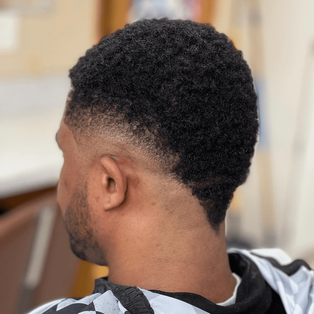 Rear view of a man with an Afro Mohawk Fade haircut. The hairstyle features natural afro-textured hair on top, styled into a Mohawk with a high fade on the sides. The fade creates a smooth gradient from the thick curls on top to the closely shaved sides. The haircut highlights the volume and texture of the afro hair while maintaining a clean and defined look. The man is wearing a black and white barber cape, sitting in a barbershop setting, showcasing the modern and stylish appearance of the Afro Mohawk Fade.