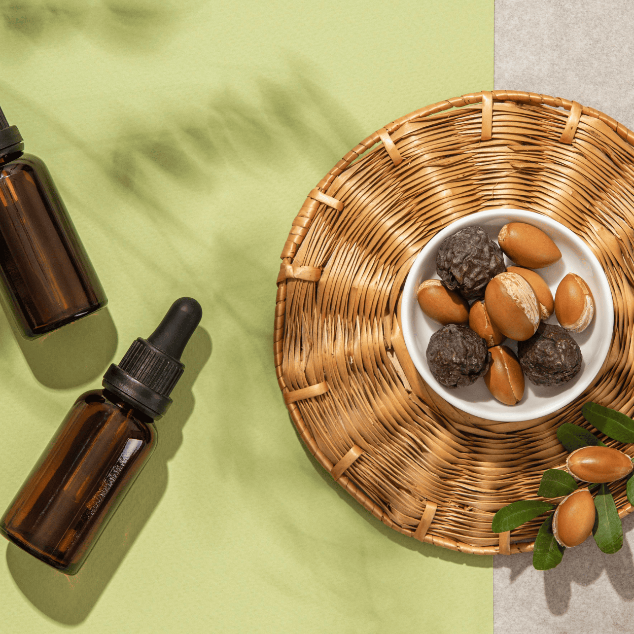 Top view of two brown dropper bottles next to a woven placemat holding a small white bowl filled with whole and cracked argan nuts on a light green and gray background, emphasizing the natural ingredients and packaging used in argan oil for beard care and grooming.