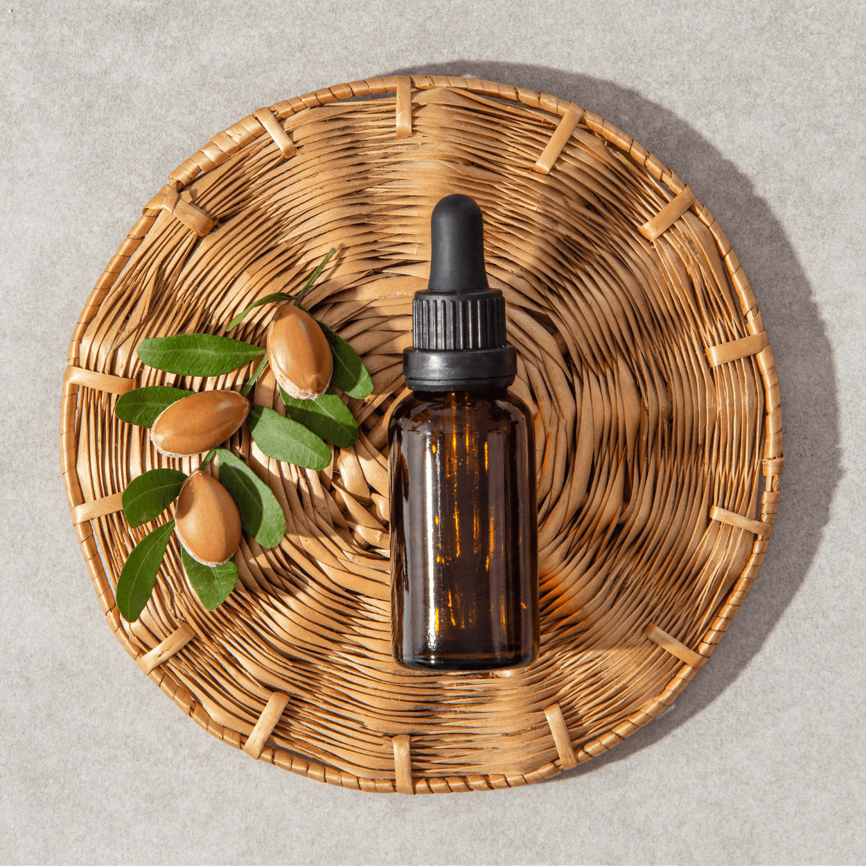 Top view of a brown dropper bottle placed on a woven placemat, accompanied by a small cluster of whole argan nuts with green leaves, set against a light gray background. This arrangement highlights the natural origins and packaging of argan oil.