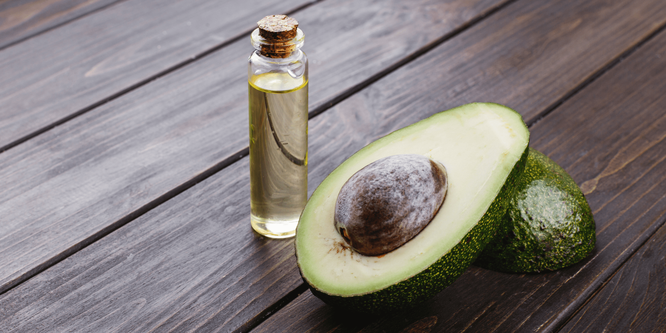 n image of a glass bottle filled with avocado oil for beard care, placed next to a fresh avocado sliced in half, revealing its green flesh and large seed, set on a rustic wooden table.