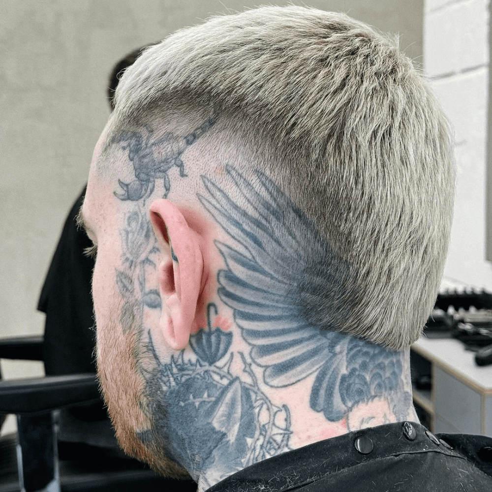 Side profile of a man with a Bald Burst Fade haircut, featuring a gradual transition from full hair on top to completely shaved sides. The hairstyle highlights the intricate tattoos covering his neck and part of his scalp, including a detailed winged design. The blonde hair color adds a modern and edgy touch to the overall look. Ideal for men seeking a bold and contemporary style.