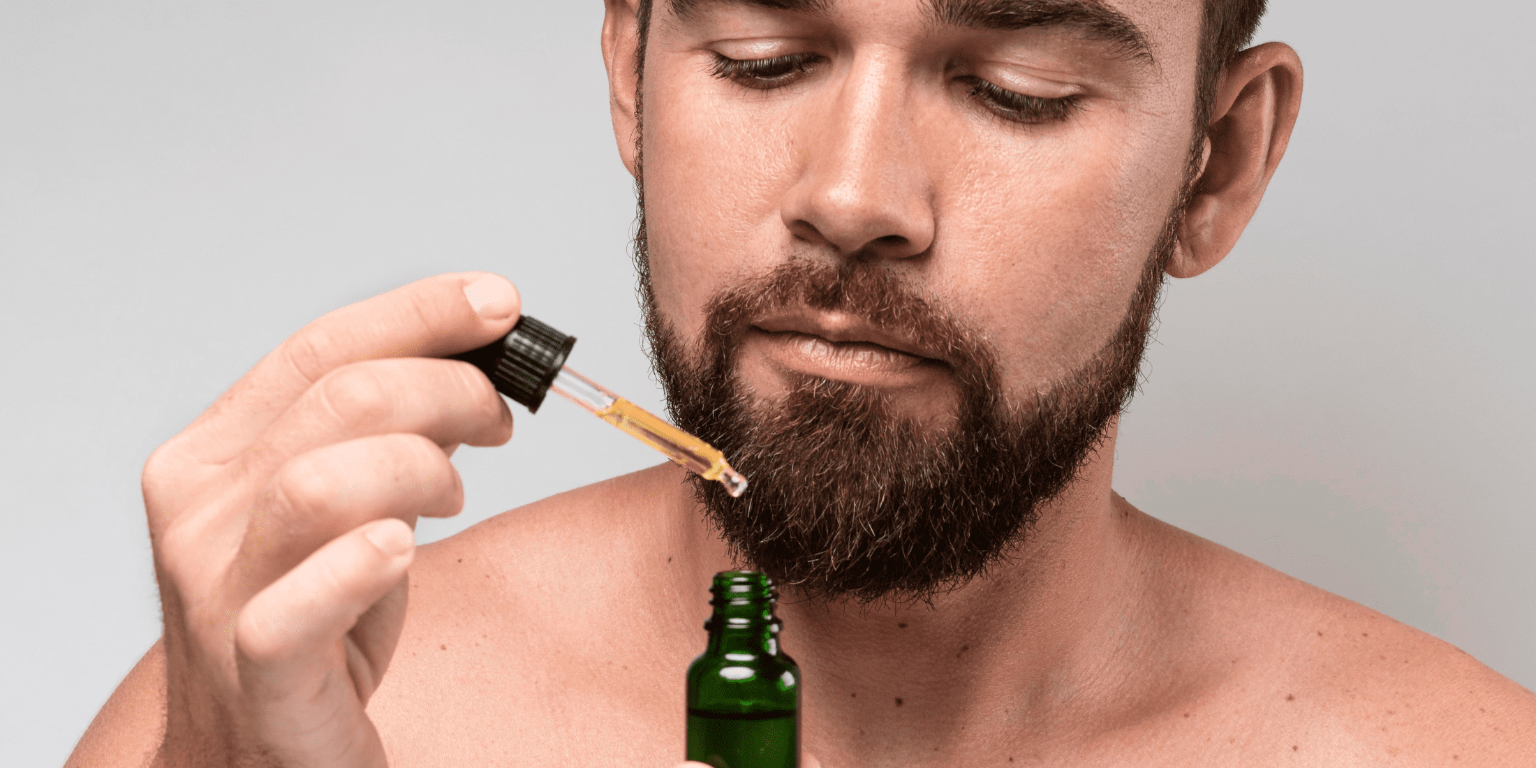 A bearded man applying avocado oil for beard care using a dropper from a green bottle. The image highlights the grooming routine and benefits of using avocado oil to maintain a healthy and well-nourished beard.