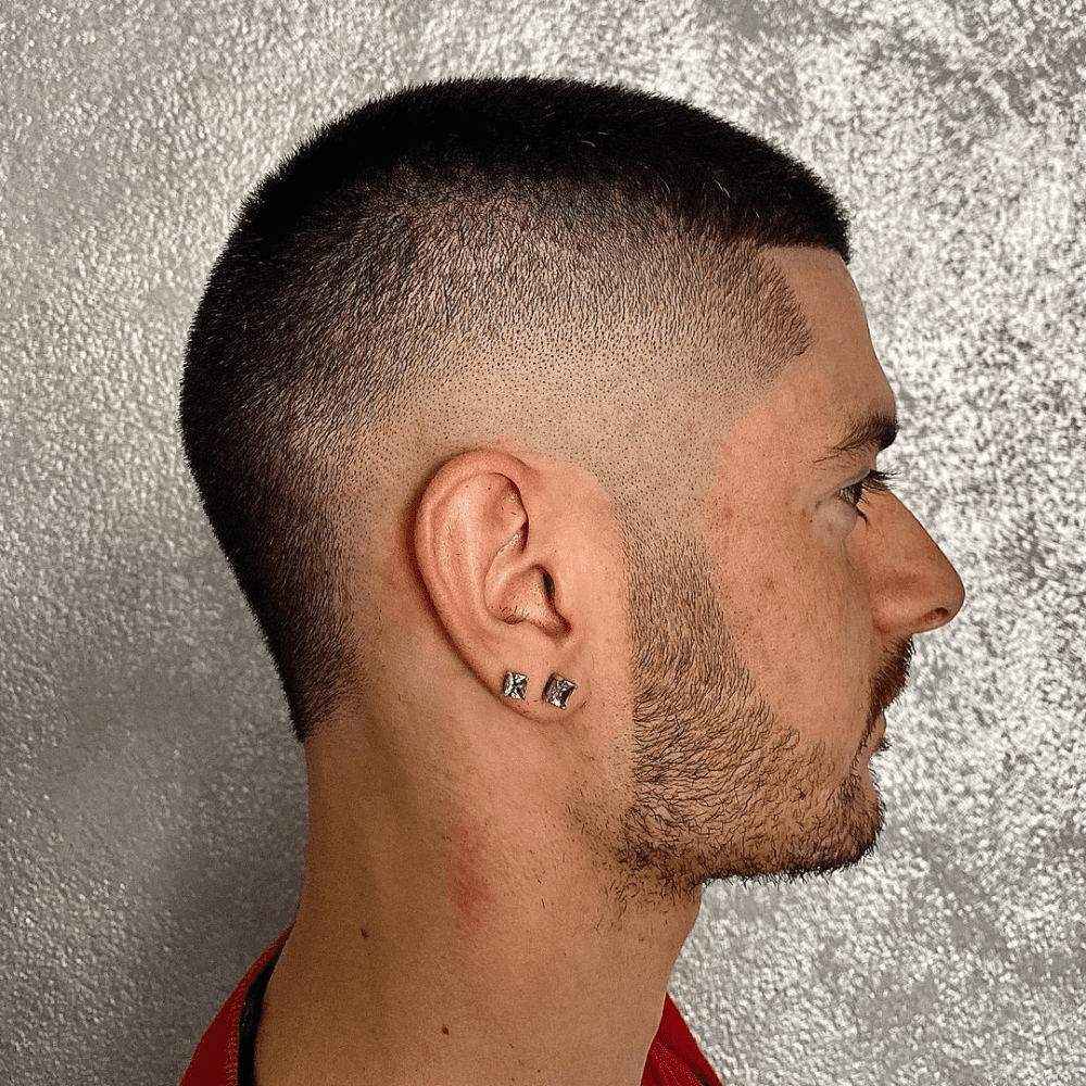 Side profile of a man with a Burst Fade Buzz Cut, showcasing a clean and low-maintenance hairstyle. The hair is uniformly short on top, blending seamlessly into a sharp fade on the sides and back. The fade begins high near the temples, creating a smooth transition that emphasizes the simplicity and precision of the buzz cut. The man has a neatly trimmed beard that complements the haircut, along with earrings that add a touch of personal style. The background is a textured, metallic silver, highlighting the sharpness and modern look of the haircut. This burst fade buzz cut is ideal for those seeking a sleek and straightforward style.