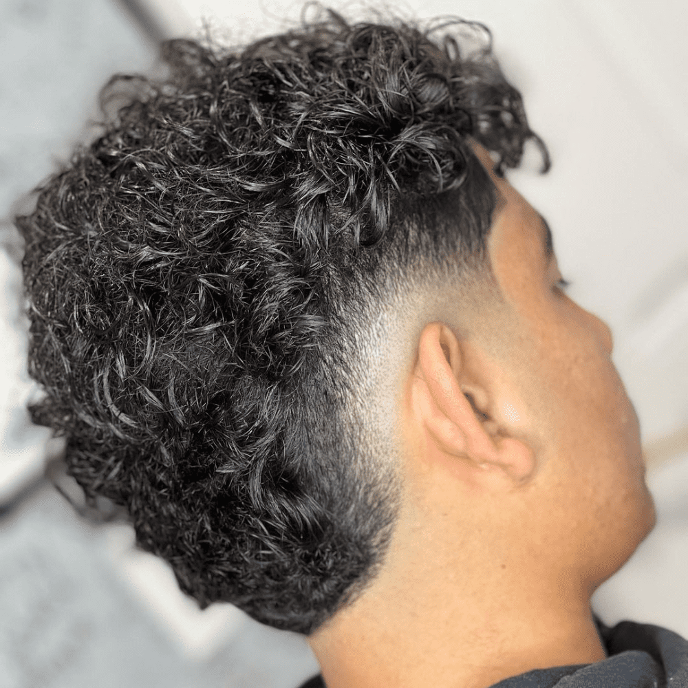 Side profile of a man with a Burst Fade haircut, featuring thick, curly hair on top that gradually fades down the sides and back. The curls are voluminous and well-defined, adding texture and movement to the hairstyle. The fade starts at the temples, creating a smooth transition that highlights the natural curls. The setting is brightly lit, drawing attention to the sharp contrast between the curly top and the clean, tapered sides. This burst fade is ideal for men with curly hair who want a stylish and modern look that enhances their natural texture.