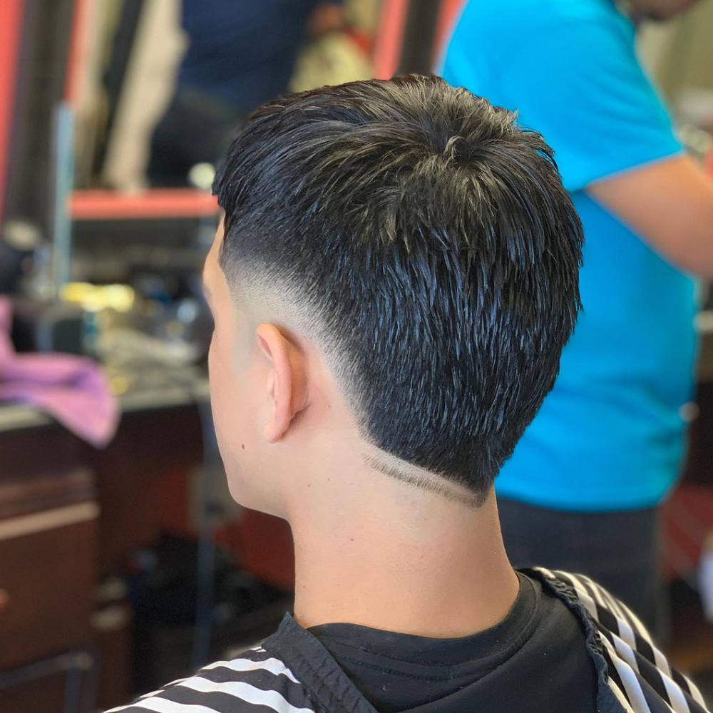 Rear view of a man with a Burst Fade Double V haircut, featuring sleek and wet-styled hair on top that smoothly transitions into a double V-shaped design at the nape of the neck. The burst fade starts high at the temples, creating a clean taper that enhances the contrast between the longer top and the closely cropped sides. The double V cut at the back adds a unique and trendy detail to the overall look. The setting is a barbershop, with the barber visible in the background, emphasizing the fresh and professionally styled haircut. This burst fade double V is ideal for men looking to make a bold and fashionable statement.