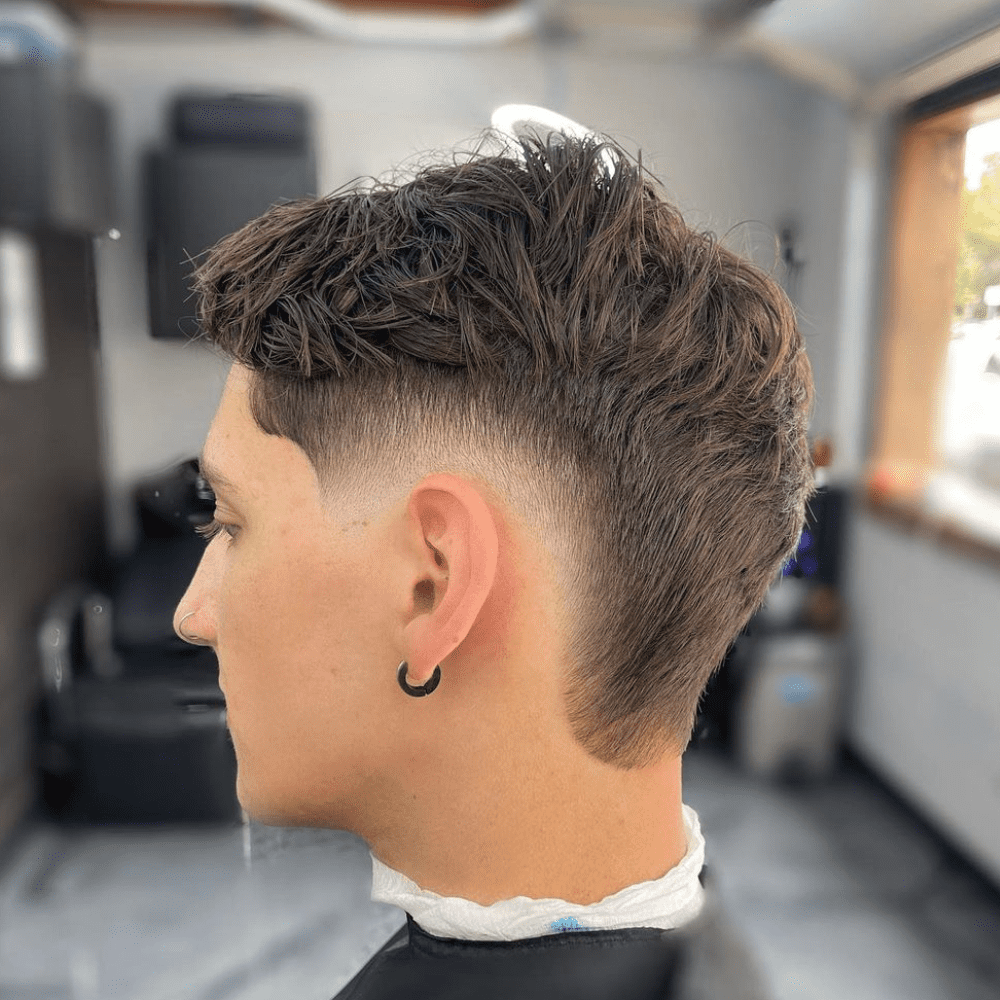 Side profile of a man with a Burst Fade haircut featuring messy, textured hair on top. The burst fade starts high near the temples and tapers down smoothly, creating a clean and stylish look that contrasts with the tousled, casual texture of the top hair. The man's hair is styled in a way that adds volume and movement, giving a relaxed and modern appearance. He is wearing a black hoop earring, adding a subtle touch of personal style. The setting is a contemporary barbershop, with a blurred background that keeps the focus on the detailed and fashionable haircut. This burst fade with messy hair is perfect for men seeking a trendy, laid-back look that is easy to maintain.