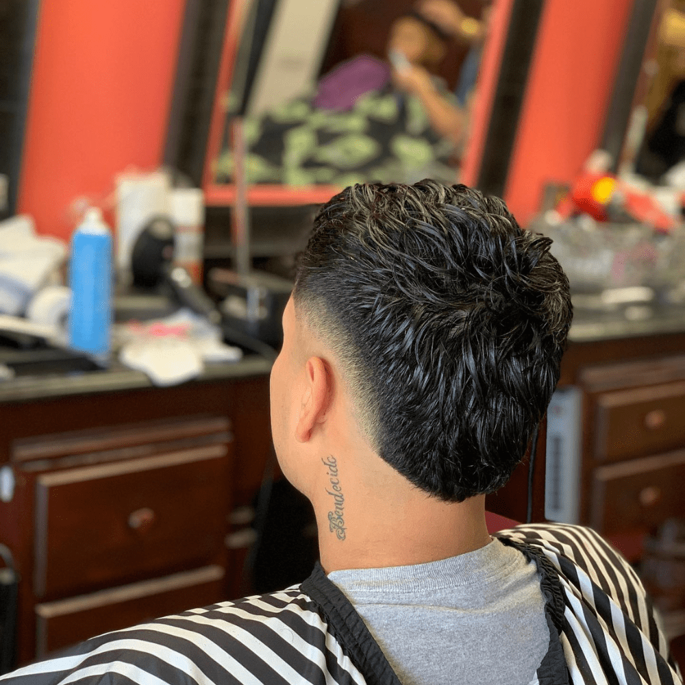 Rear view of a man with a Burst Fade V Cut haircut, showcasing sleek and wet-styled hair on top that transitions smoothly into a sharp V-shaped cut at the nape. The burst fade starts high at the temples and tapers down to the V cut, creating a clean and stylish appearance. The hair is dark and glossy, adding a polished finish to the look. The man has a tattoo on the side of his neck, adding a personal touch to the overall style. The setting is a modern barbershop, with a blurred background that emphasizes the haircut's precision and detail. This burst fade V cut is ideal for men seeking a trendy and distinctive hairstyle.