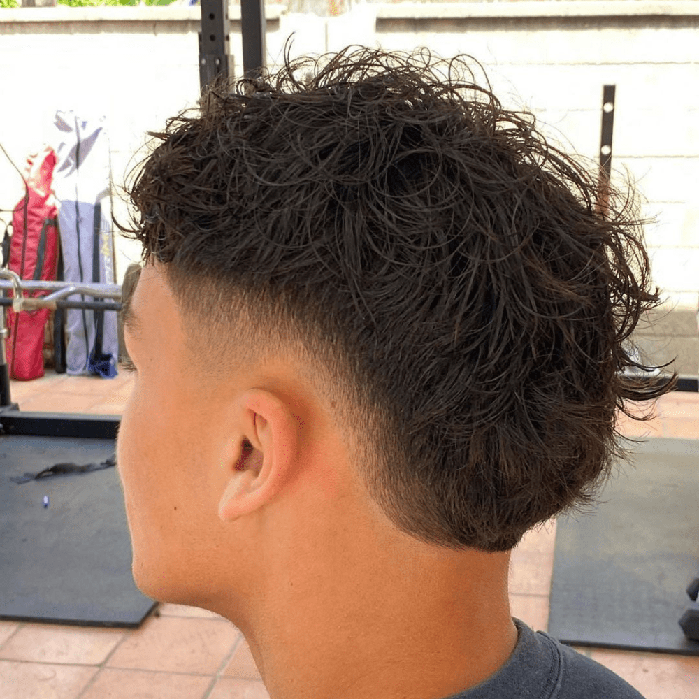 Side profile of a man with a Burst Fade haircut, featuring wavy hair on top that gradually transitions into a fade on the sides and back. The waves are textured and voluminous, adding a natural flow to the hairstyle. The fade starts at the temples, creating a smooth and clean taper that enhances the wavy texture. The setting is outdoors, with gym equipment in the background, highlighting the fresh and stylish haircut. This burst fade is perfect for men with wavy hair who want a modern and effortlessly cool look.