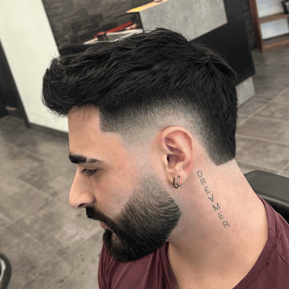 Side profile of a man with a Burst Fade haircut complemented by a full beard. The hair on top is styled with volume and texture, while the burst fade starts high at the temples and tapers down smoothly, creating a clean and sharp look. The beard is neatly trimmed and well-groomed, blending seamlessly with the fade to create a cohesive and stylish appearance. The man has a tattoo on the side of his neck that reads "DREAMER," adding a personal touch to his look. The setting is a modern barbershop, emphasizing the fresh and professional styling of the haircut and beard. This burst fade with beard is ideal for men looking to achieve a trendy and polished appearance.