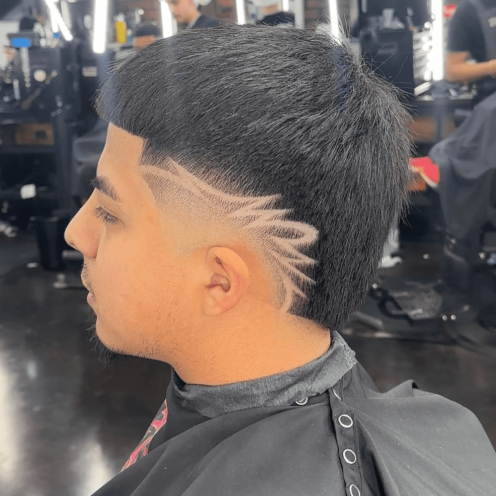 Side profile of a man with a Burst Fade haircut featuring a creative design shaved into the sides. The haircut has a neat and textured top, while the burst fade starts high near the temples and gradually tapers down. The artistic design adds a unique and eye-catching element, showcasing intricate lines and patterns that enhance the overall style. The setting is a modern barbershop, with other clients and barbers visible in the background, emphasizing the professional and trendy atmosphere. This burst fade with design is perfect for men looking to express their individuality and stand out with a stylish and customized haircut.