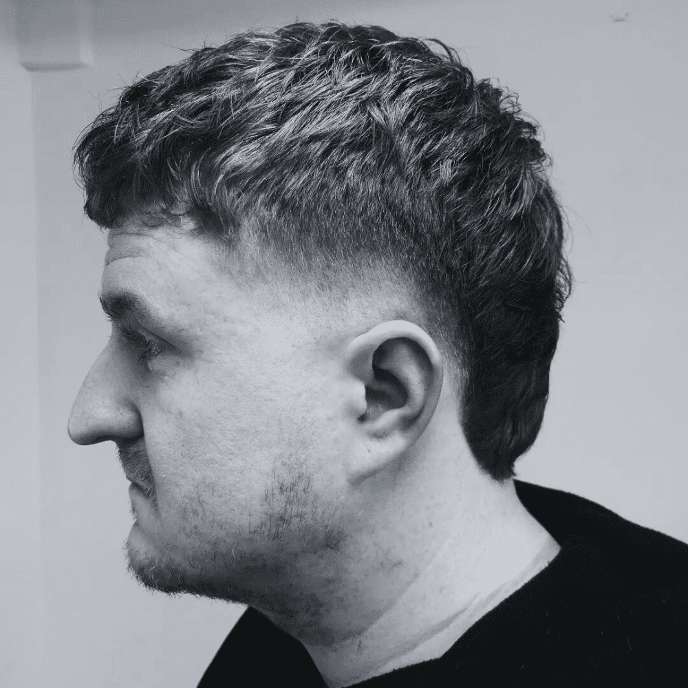 Side profile of a man with a Burst Taper Fade haircut, featuring short, textured hair on top that gradually tapers down the sides and back. The fade starts at the temples and blends seamlessly into the natural hairline, creating a clean and polished look. The hairstyle is slightly longer on top, allowing for a subtle texture that adds volume and movement. The man has a neatly trimmed beard, enhancing the overall well-groomed appearance. The setting is a minimalist indoor background, emphasizing the sharp and precise nature of the haircut. This burst taper fade is ideal for men seeking a refined and stylish look that is easy to maintain.