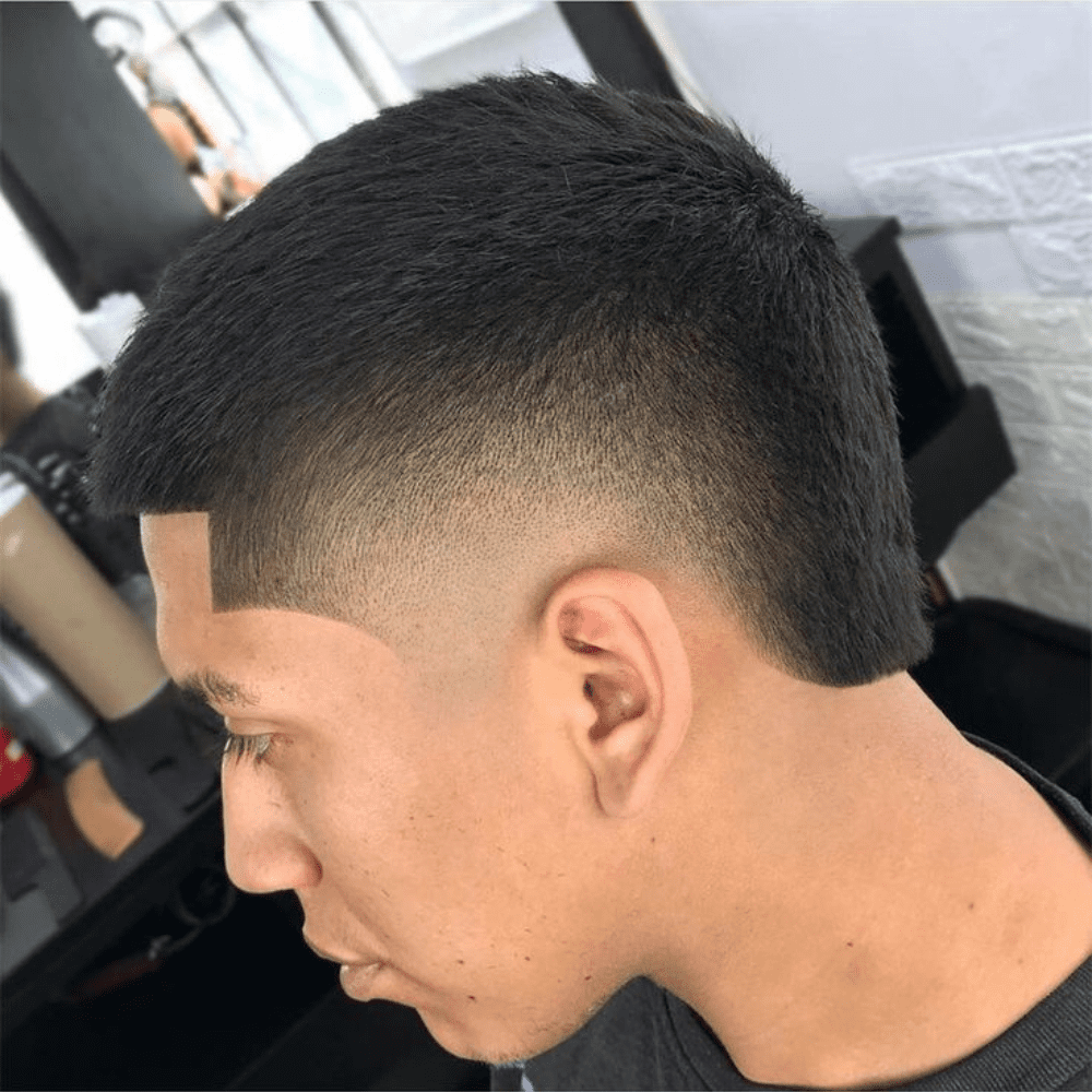 A side profile of a man with a buzz cut faux hawk hairstyle. The haircut features short, textured hair on top that gradually tapers down to a fade on the sides and back. The top is styled slightly longer in the center, mimicking the shape of a faux hawk without the extreme length, creating a subtle yet edgy look. The hairline is sharply defined with a clean, angular line-up at the forehead, adding precision to the style. The overall appearance is modern and stylish, offering a balanced blend of boldness and simplicity. The setting appears to be a barbershop, with grooming products visible in the background.