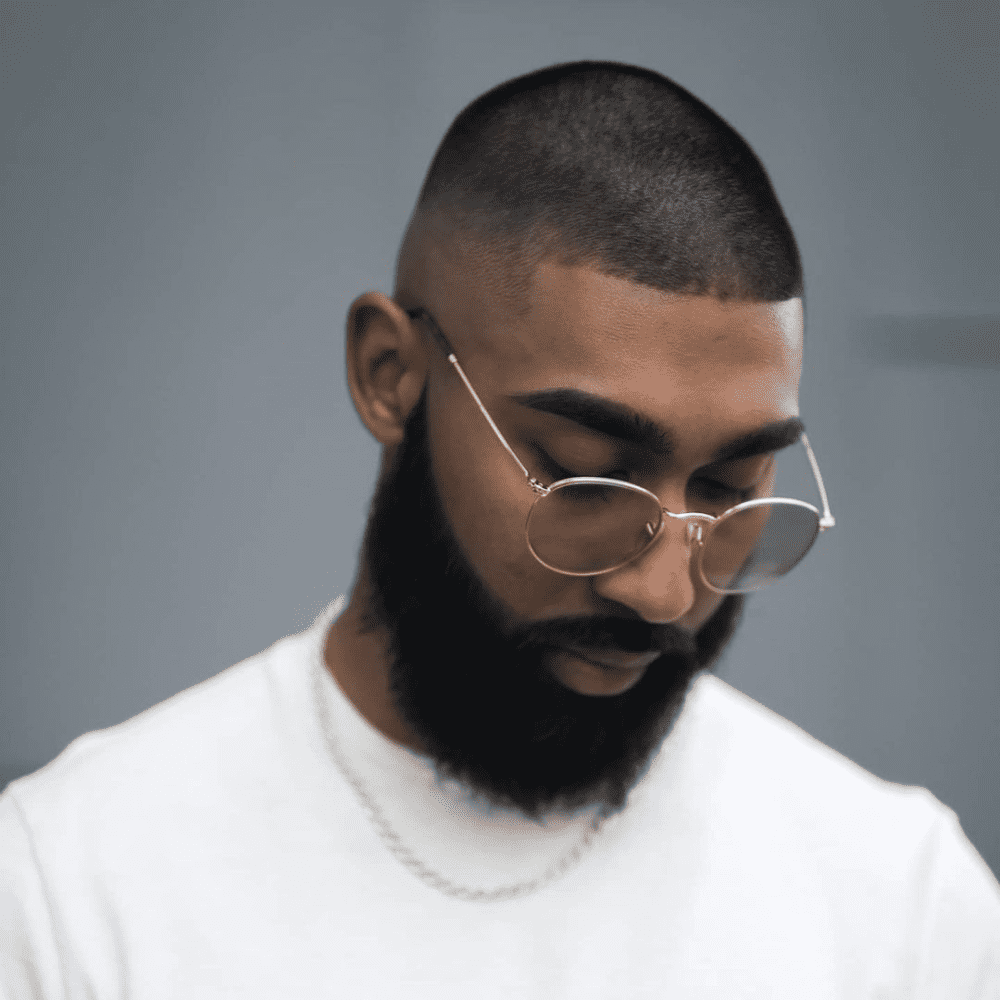 Front profile of a man with a buzz cut high fade haircut, featuring very short hair all around with a high fade on the sides, creating a clean and minimalist look. The man is wearing glasses and has a full, well-groomed beard, enhancing his overall appearance with a blend of simplicity and style.