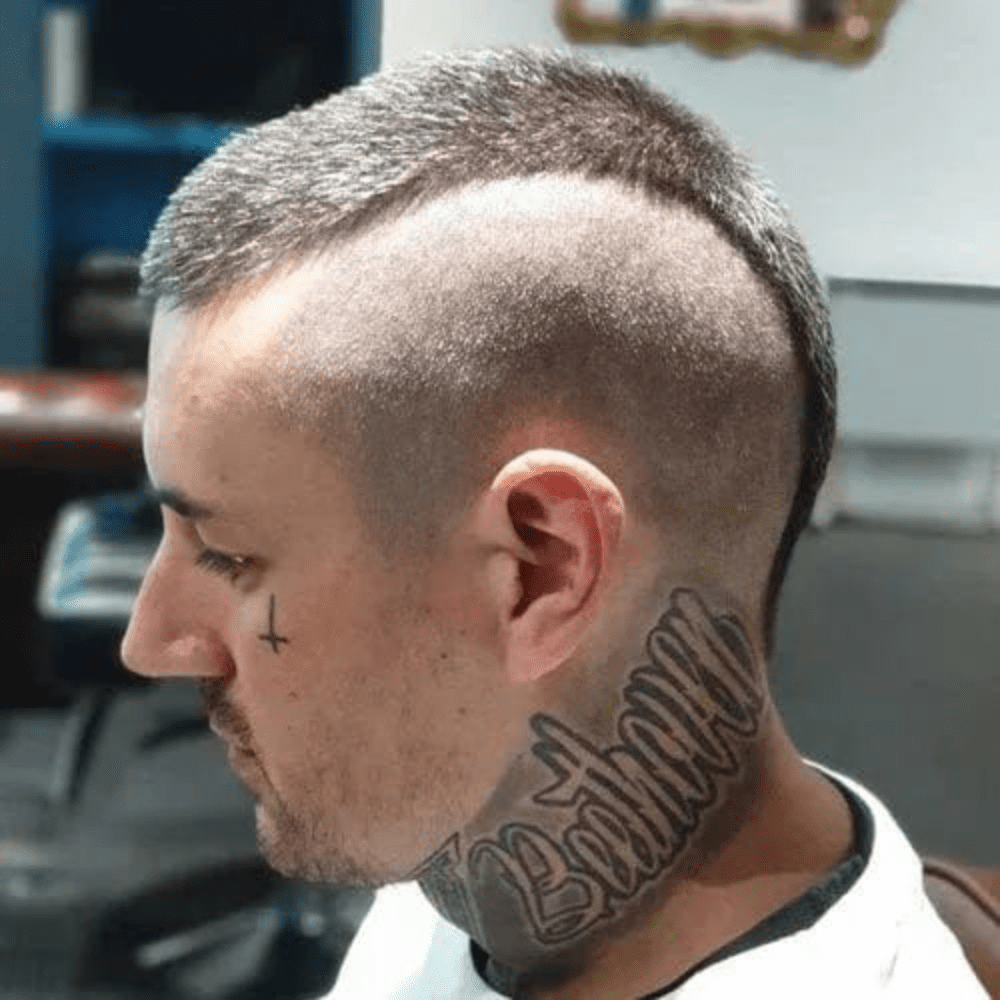 A side profile of a man with a buzz cut mohawk hairstyle. The haircut features very short, evenly trimmed hair on the sides with a narrow strip of longer hair running from the front to the back of the head, creating a mohawk. The sides are shaved down to almost the skin, emphasizing the contrast between the mohawk and the buzzed sides. The man has several prominent tattoos, including a large script tattoo on his neck and a small cross tattoo on his cheek, adding to his bold and edgy appearance. The overall look is striking and unconventional, perfect for someone seeking a rebellious and distinctive style. The background appears to be a barbershop setting.