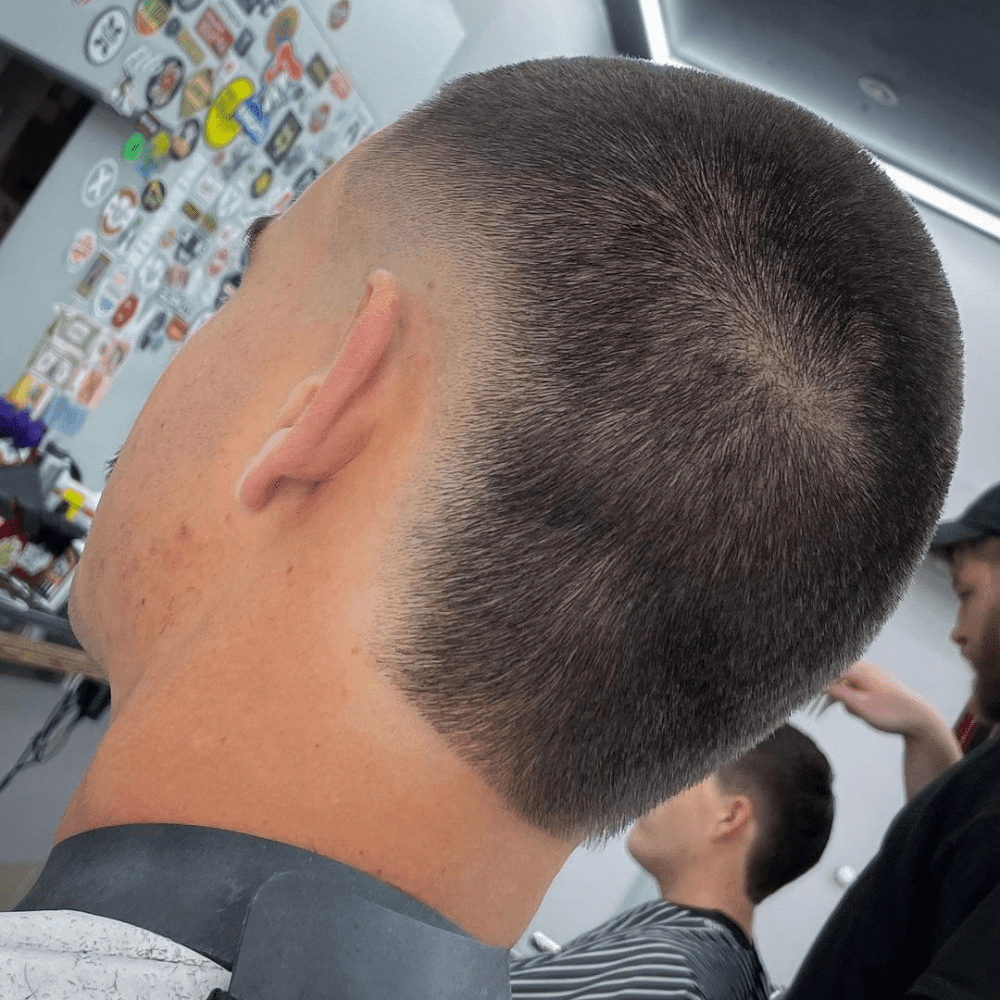 Rear view of a man with a Buzz Cut Mohawk Fade haircut. The hairstyle features a short, uniform buzz cut on top with a high fade on the sides and back, creating a subtle Mohawk effect. The high fade adds a clean and sharp contrast to the closely cropped hair, giving a modern and low-maintenance look. The man is seated in a barbershop, with a decorated wall and another client in the background. The Buzz Cut Mohawk Fade combines simplicity with a touch of edginess, perfect for a neat and stylish appearance.