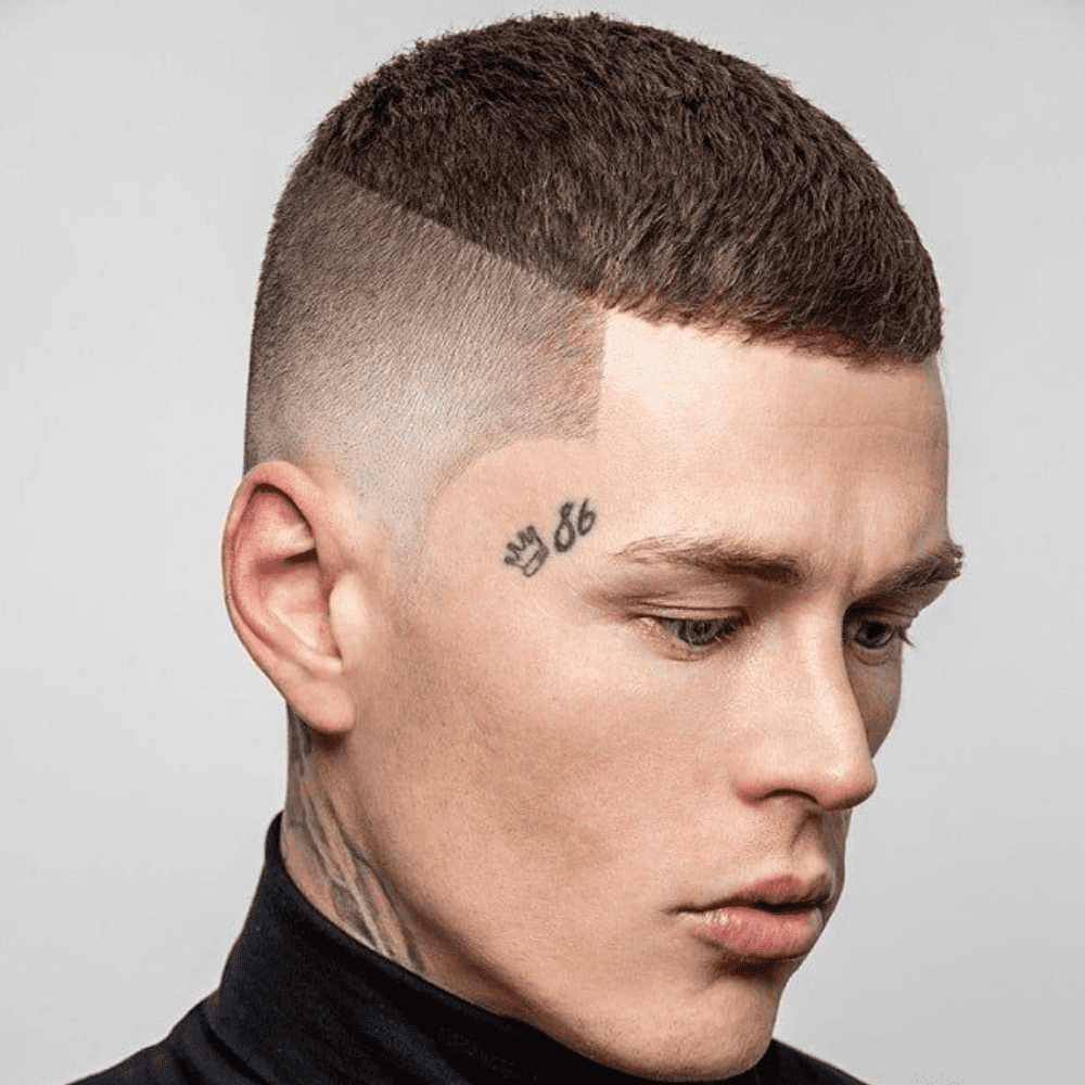 A close-up side profile of a man with a buzz cut featuring a disconnected undercut. The haircut showcases short, textured hair on top, sharply contrasted by the disconnected undercut on the sides, which is closely shaved down to the skin. The hairline is clean and angular, adding a precise and defined look to the style.