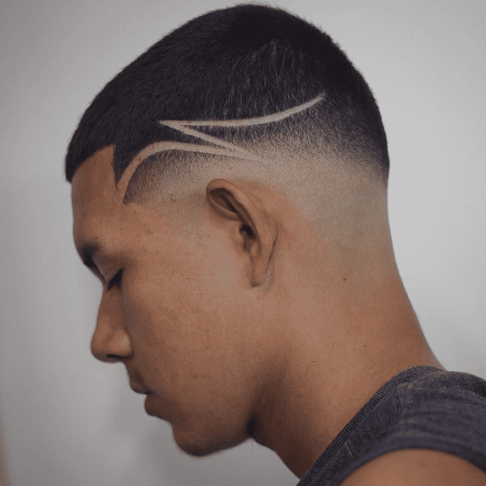 A side profile of a man with a buzz cut featuring a creative design. The haircut is short and uniform on top, with a smooth fade down the sides and back. A striking design is shaved into the faded section, consisting of clean, curved lines that sweep from the temple area towards the back of the head. The design adds a dynamic and artistic element to the traditional buzz cut, making it a unique and eye-catching style. The man’s head is slightly tilted down, highlighting the intricate detail of the shaved pattern. The overall look is bold, modern, and perfect for someone looking to make a fashion statement with their haircut.