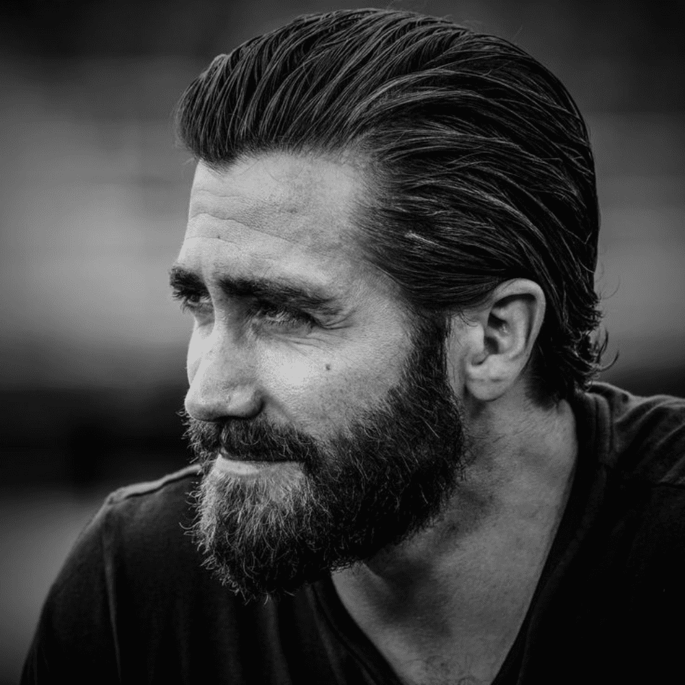 Black and white portrait of a man with a classic slicked back haircut and a thick beard, wearing a dark shirt. This image exemplifies a timeless, sophisticated slicked back hairstyle, perfect for men seeking a polished and masculine look in 2024.