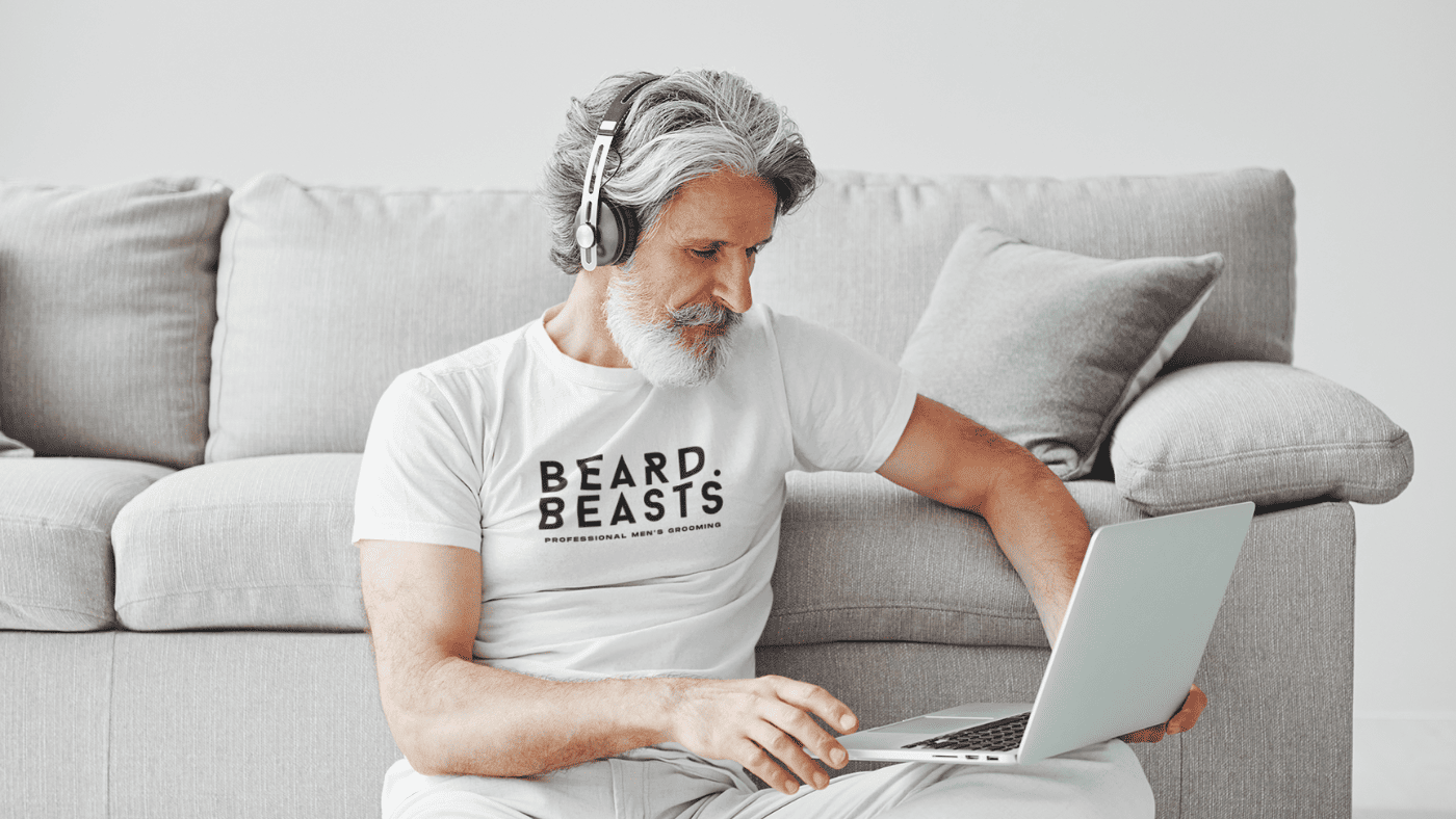 A stylish man with a gray beard wearing a white Beard Beasts t-shirt, sitting comfortably on the floor using a laptop with headphones, representing a modern and approachable lifestyle.