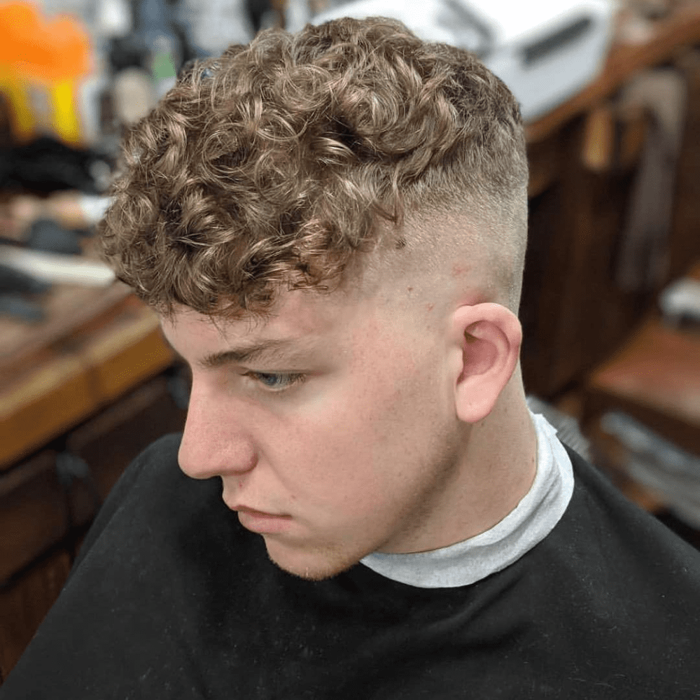 This image displays a stylish Textured Crop Curls haircut for men, featuring short sides and back with slightly longer, textured curls on top. The clean fade on the sides provides a neat contrast to the voluminous and defined curls, creating an effortlessly chic look. Perfect for men who want a low-maintenance yet fashionable style, this haircut enhances the natural curl pattern while keeping the overall appearance polished. The textured crop curls are a trendy choice for 2024, ideal for various face shapes and easy to style with minimal products.