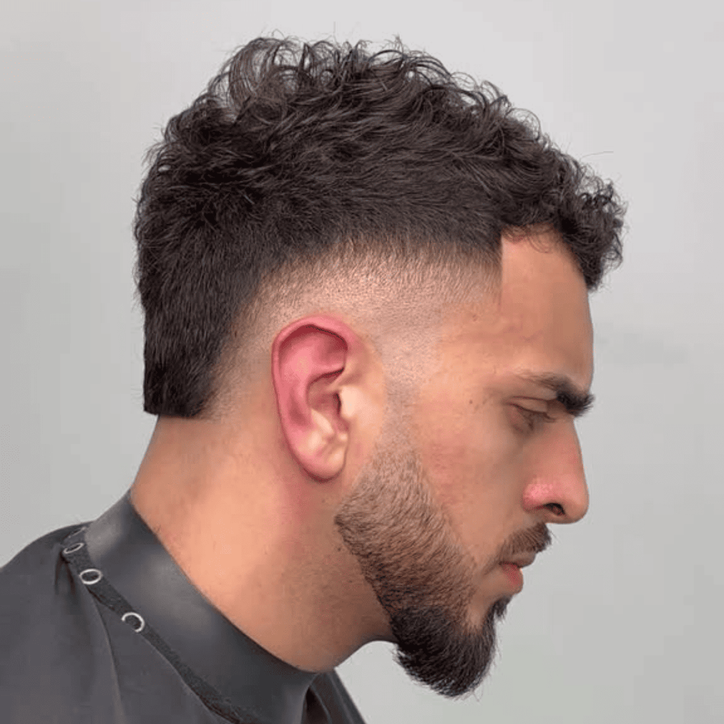 Side profile of a man with a curly haircut featuring a burst fade around the ears. The voluminous curls on top contrast with the clean, tapered fade, while a well-groomed beard adds definition to the overall look.