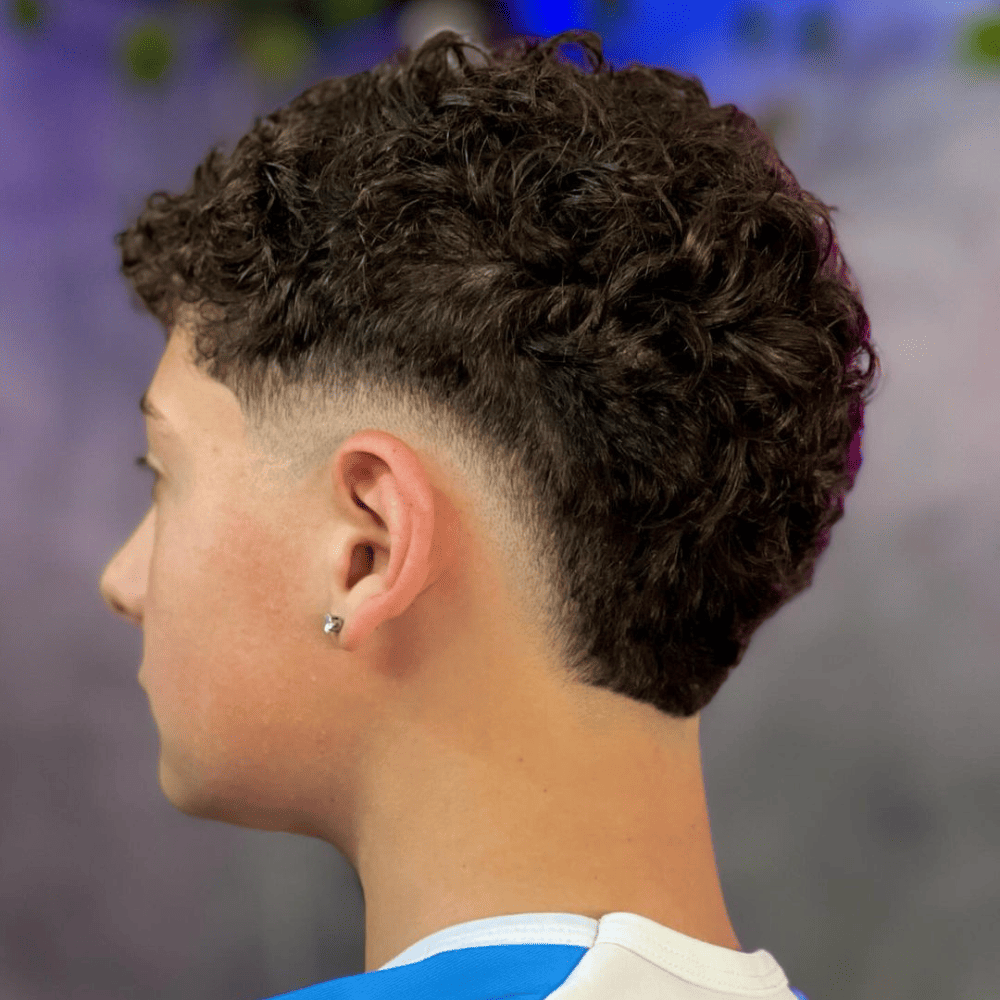 A modern Curly Burst Fade haircut for men. The image features a young man with tightly coiled curls on top, gradually transitioning into a burst fade around the ears and down to the nape of the neck. This haircut creates a striking contrast between the voluminous curls and the closely shaved sides, enhancing the natural texture of the curly hair. The back view emphasizes the precise fade and the stylish blend, perfect for those looking for trendy short curly haircuts for men.