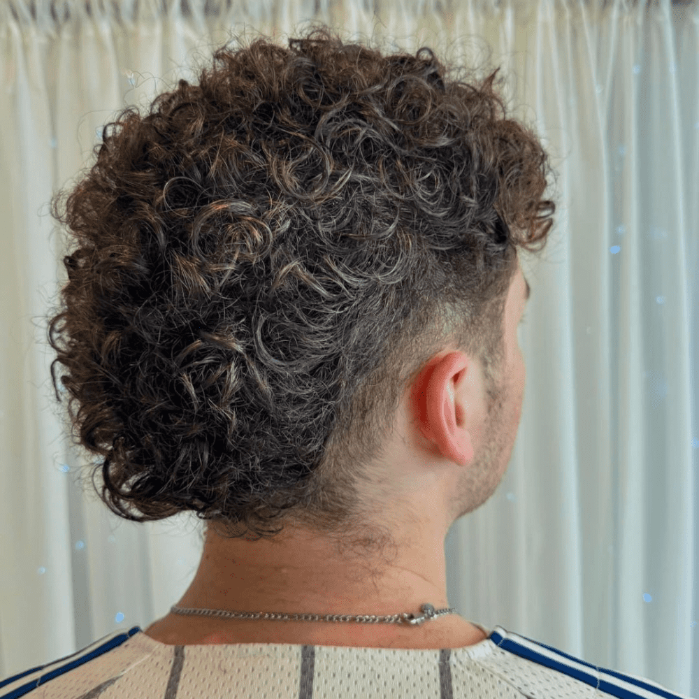 This image features a distinctive Curly Faux Hawk Mullet haircut for men, combining the edgy elements of a faux hawk with the length and texture of a mullet. The hairstyle showcases voluminous curls that run from the top to the back, with the sides closely shaved or faded, emphasizing the natural curl pattern. Perfect for men who want a unique and bold look, the curly faux hawk mullet offers a blend of modern and retro styles. This haircut is ideal for various face shapes and is a trendy choice for 2024, highlighting creativity and individuality.