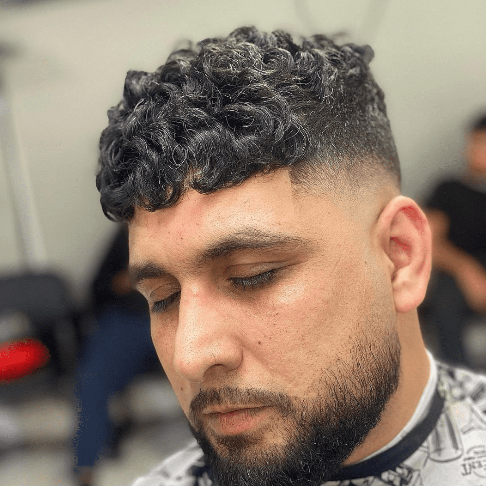 Close-up of a man with a curly high fade haircut, featuring tightly curled hair on top that transitions smoothly into closely shaved sides. The high fade creates a sharp contrast, highlighting the natural texture of the curls. The look is complemented by a short, well-groomed beard, giving a stylish and modern appearance. The setting appears to be a barber shop, enhancing the professional grooming atmosphere.