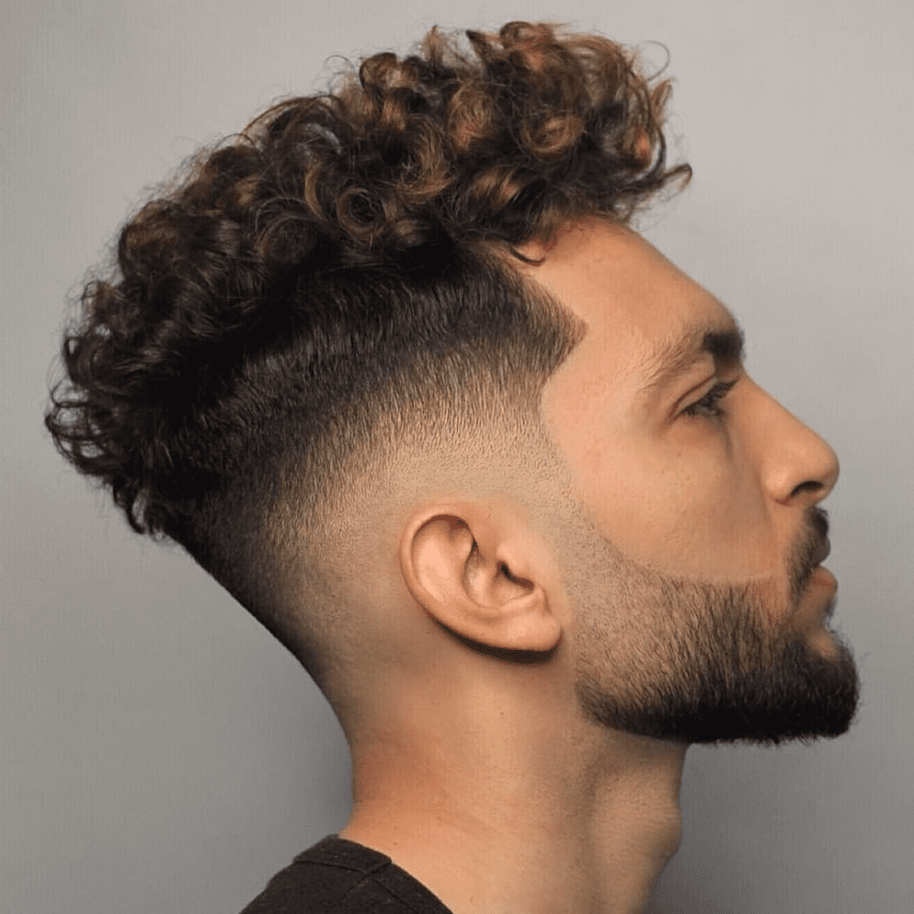 This image features a striking Curly High Top Fade haircut for men, showcasing voluminous curls on the top with a sharp, high fade on the sides and back. This trendy hairstyle emphasizes the natural texture of curly hair while maintaining a clean and edgy appearance. Ideal for men looking to combine classic and contemporary styles, the curly high top fade suits various face shapes and provides a fresh, modern look. This haircut is a popular choice for 2024, offering both style and ease of maintenance.