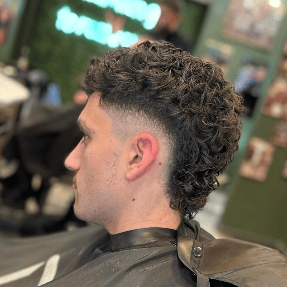 Close-up of a man with a Curly Mohawk Fade haircut. The hairstyle features natural curls on top, gradually fading into shorter hair on the sides. The haircut highlights the texture and volume of the curly hair, with a clean, sharp fade that emphasizes the contrast between the top and sides. The man is seated in a barber chair in a modern barbershop.