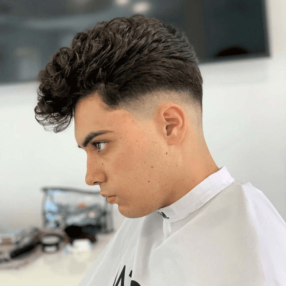 A young man is pictured in a barbershop, featuring a modern curly pompadour haircut. His hair is voluminous and curly on top, styled in a high pompadour, while the sides and back are neatly tapered with a fade. The contrast between the curly top and the clean, faded sides creates a striking, contemporary look. The barbershop environment in the background appears well-lit and professional, enhancing the focus on the stylish haircut. The man's expression is serious, emphasizing the sophistication of his new hairstyle.