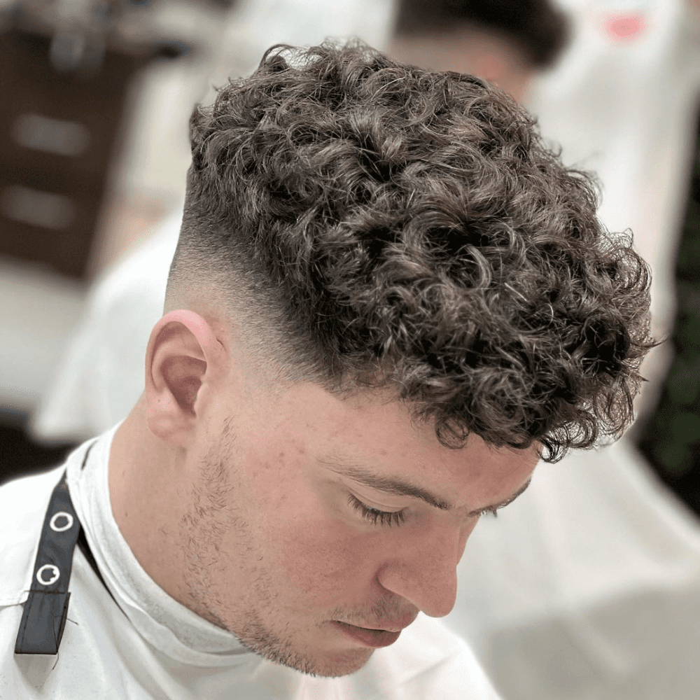 A stylish Curly Skin Fade haircut for men. The image showcases a young man with thick, natural curls on top, gradually fading into a clean-shaven skin fade on the sides and back. This modern haircut emphasizes the natural texture and volume of curly hair, creating a sharp and edgy look. The close-up view highlights the precise fade and well-defined curls, offering inspiration for those seeking trendy curly haircuts for men.