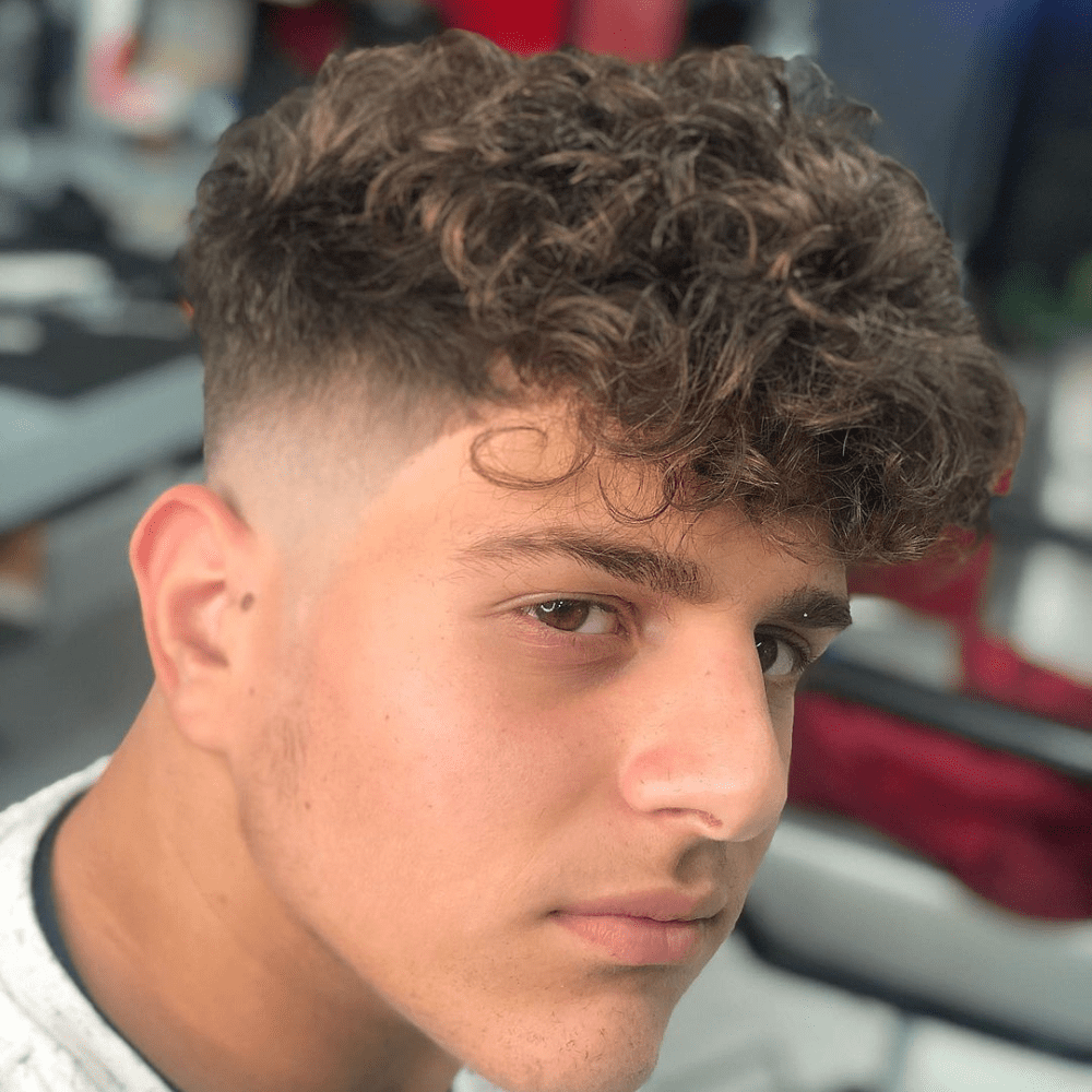 This image features a trendy curly undercut hairstyle for men, showcasing voluminous curls on top with a sharp, high fade on the sides and back. This modern haircut accentuates the natural texture of curly hair, providing a stylish and clean look. Perfect for men who prefer a low-maintenance yet fashionable style, this curly undercut is ideal for various face shapes and offers a fresh, contemporary appeal. This haircut is a popular choice for those looking to embrace their curls while keeping a neat and edgy appearance in 2024.