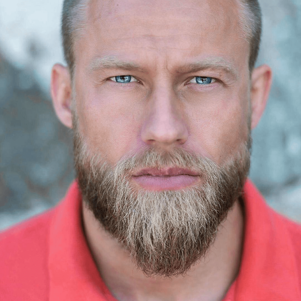 Close-up of a man with a well-groomed ducktail beard, showcasing a tapered shape that enhances his strong jawline, set against a blurred outdoor background, highlighting the beard's texture and style.