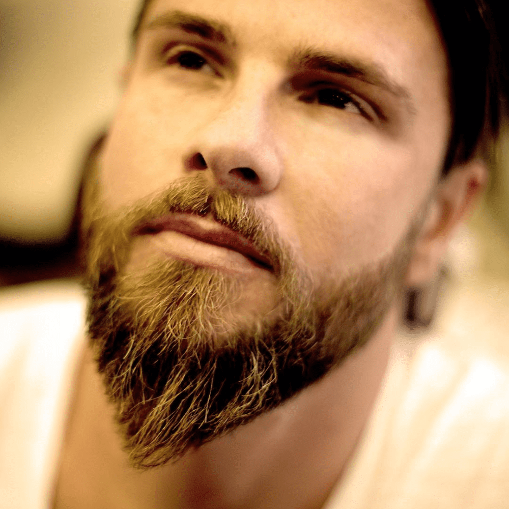 A close-up of a man with a well-groomed Ducktail beard, a popular medium beard style characterized by a fuller chin area that tapers down to a point, resembling a duck's tail. The Ducktail beard enhances his facial features, providing a stylish and distinctive look that combines fullness with a unique shape. This medium beard style is ideal for those seeking a bold and polished appearance, perfect for 2024 trends. The Ducktail beard offers a sophisticated yet rugged option for men aiming to make a statement with their facial hair.