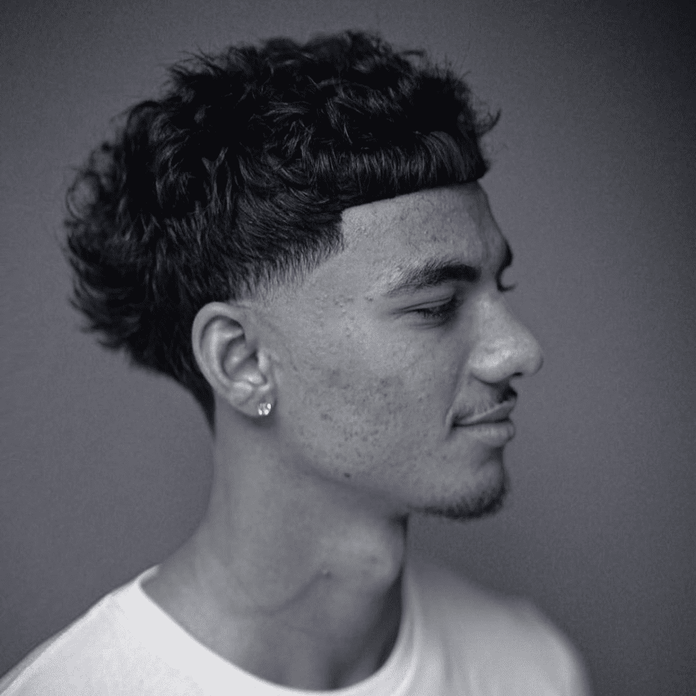 Side profile of a young man with an Edgar Cut Taper Fade haircut, featuring a short, blunt fringe with textured hair on top and a clean taper fade on the sides and back. The hairstyle highlights a bold and trendy look, perfect for men seeking unique fade haircuts in 2024. The defined fringe and clean lines enhance the overall edgy appearance