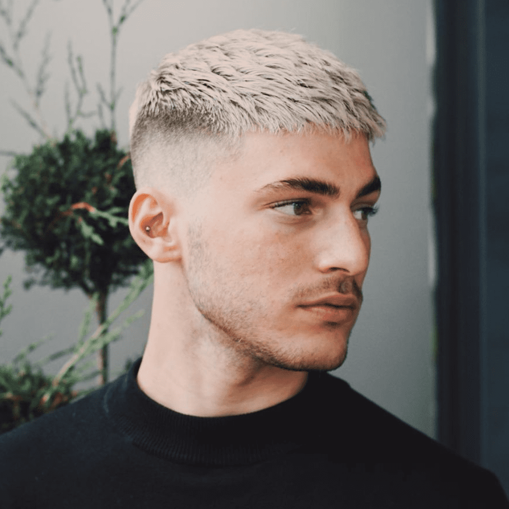 Side profile of a man with a French crop high fade haircut, featuring short, textured hair on top with a straight fringe. The sides are faded high, creating a sharp contrast that enhances the clean, modern look. The hair is dyed a platinum blonde, adding a unique and stylish touch to the overall appearance.