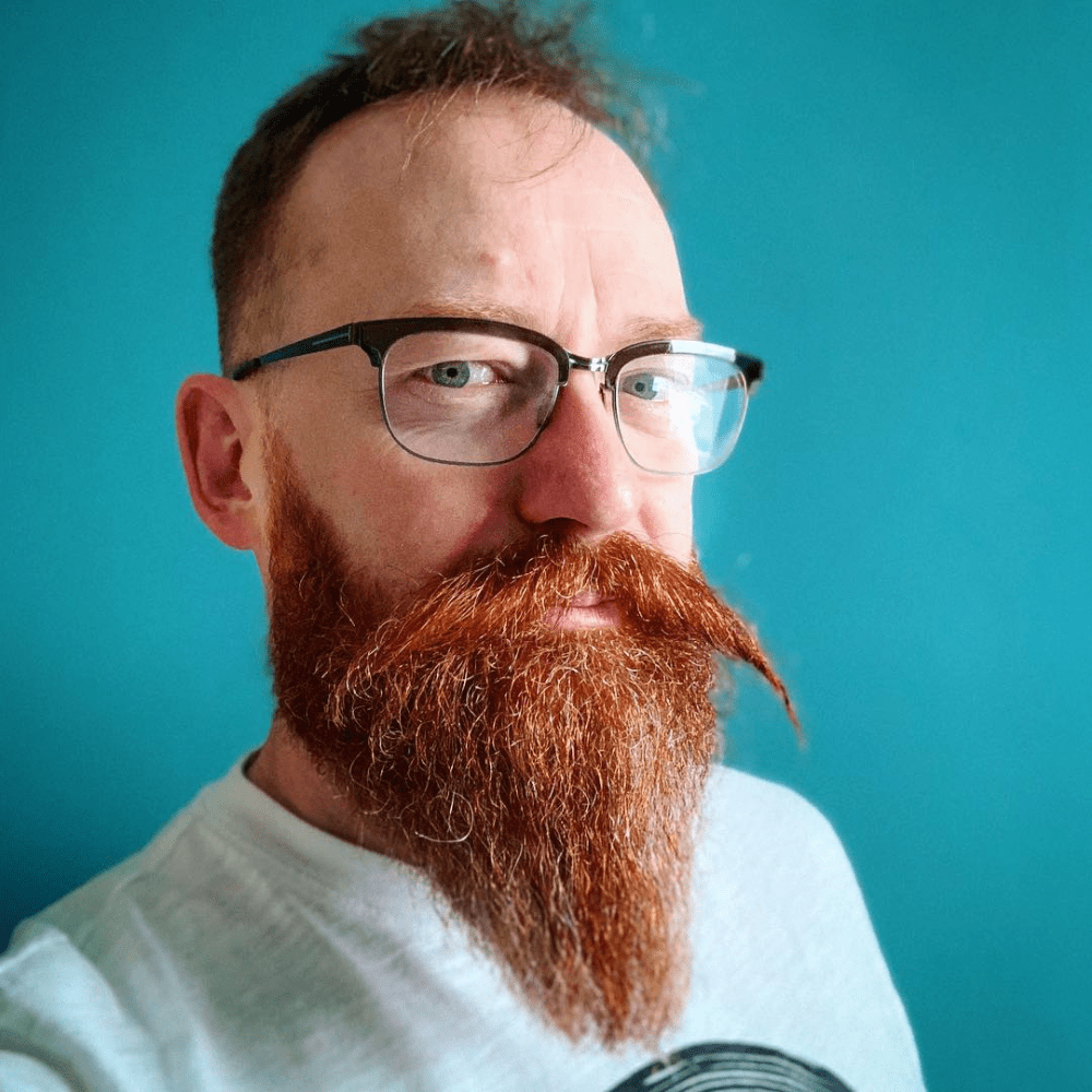 Portrait of a man with a vibrant red, full ducktail beard and styled mustache, wearing glasses and a light-colored shirt, set against a teal background, highlighting the beard's fullness and meticulous grooming.