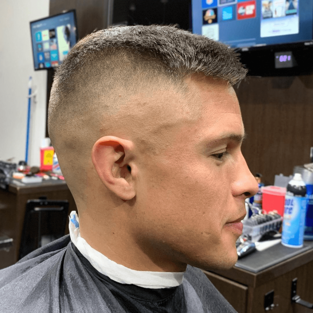 Side profile of a man with a high and tight haircut, featuring very short hair on top and closely shaved sides. The haircut showcases a military-inspired style with a clean and disciplined look. The setting appears to be a barber shop, with grooming tools and products visible in the background.