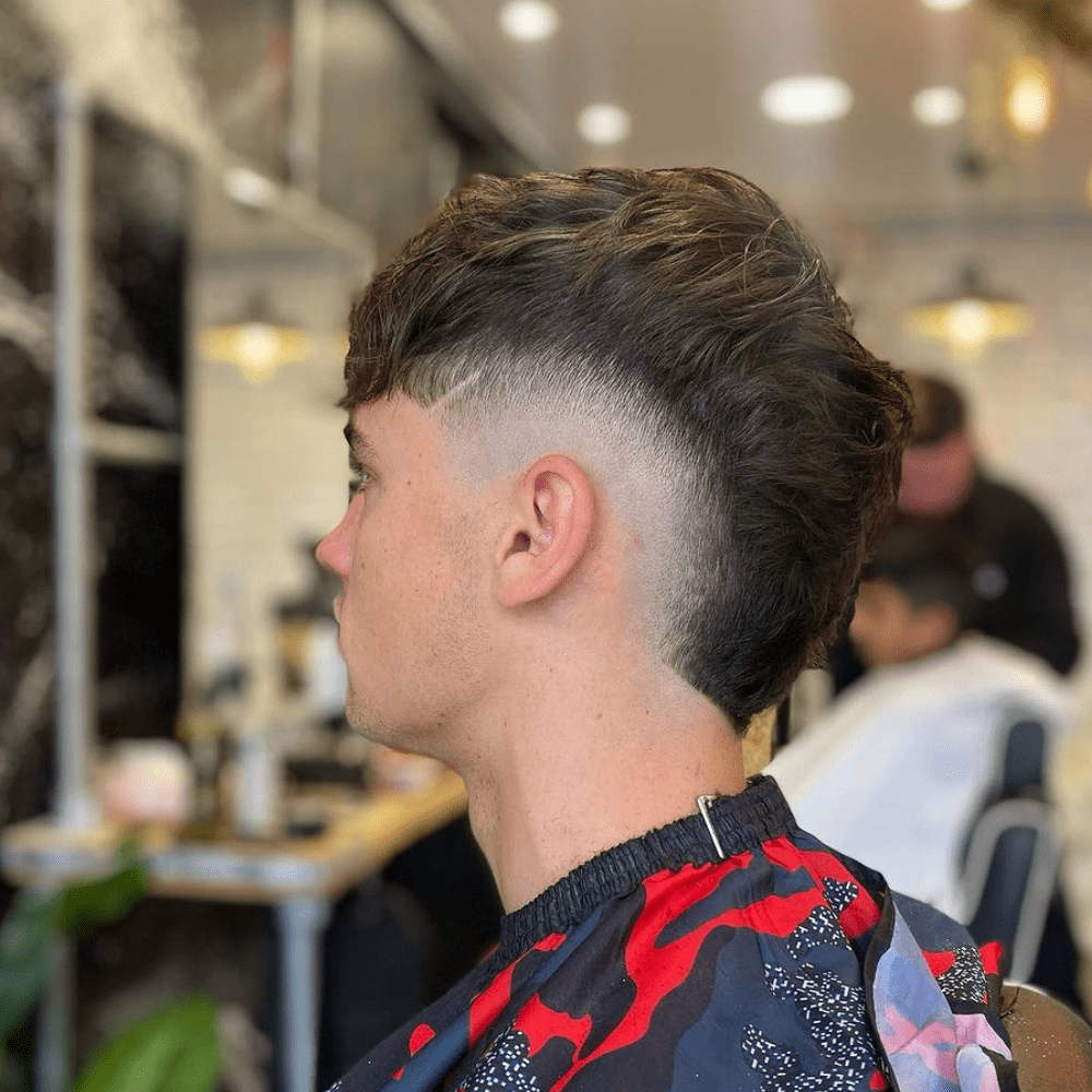 Side profile of a man with a High Burst Fade haircut, featuring a bold and dramatic fade that starts high above the temples. The hair on top is longer and styled with texture, while the sides and back are closely cropped, creating a striking contrast. The high burst fade accentuates the volume and length on top, providing a modern and edgy appearance. The setting is a contemporary barbershop, with a blurred background that emphasizes the sharp and clean lines of the haircut. This high burst fade is perfect for those looking to make a strong style statement.