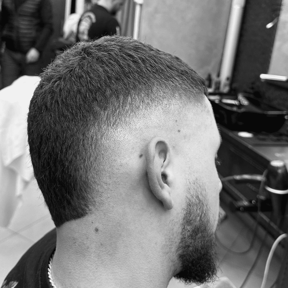 Side profile of a man with a high burst fade haircut, showcasing the unique rounded fade around the ear that transitions smoothly from the longer hair on top to the closely shaved sides, accompanied by a short, trimmed beard.