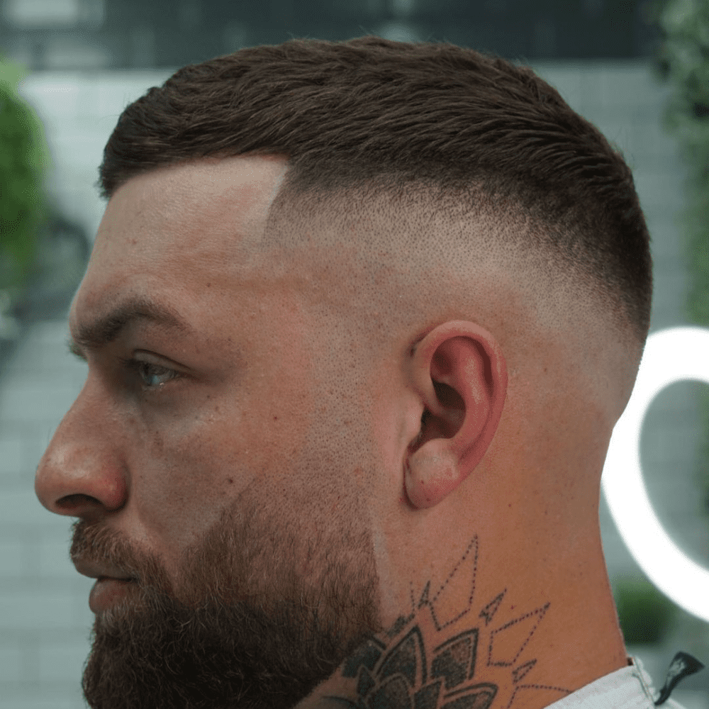 Side profile of a man with a high drop fade haircut, showing the gradual fade that drops behind the ear, blending seamlessly from the longer hair on top to the closely shaved sides. The look is completed with a well-groomed beard and a visible neck tattoo.