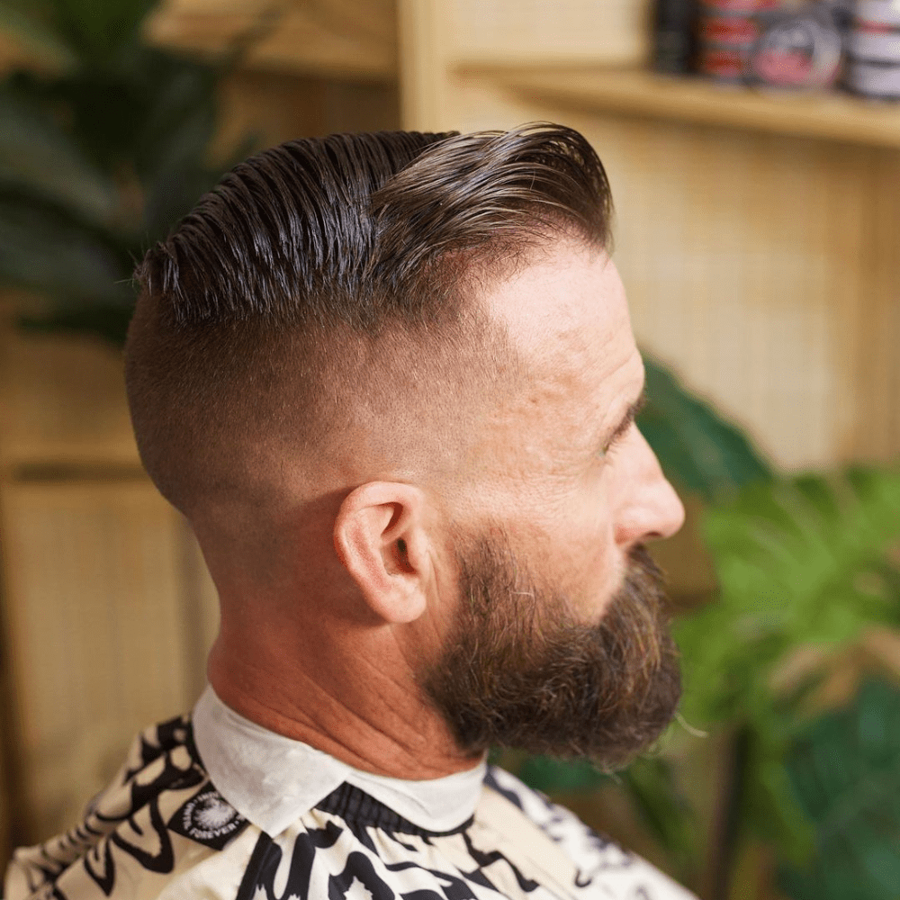 Side profile of a man with a high fade comb over haircut, featuring longer hair on top that is neatly combed over to one side. The sides are closely shaved with a high fade, creating a sharp contrast. The look is complemented by a full, well-groomed beard. The setting appears to be a barber shop, with grooming products and plants in the background, enhancing the stylish and professional appearance.