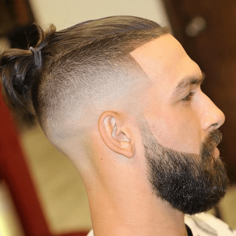 Side profile of a man with a high fade haircut featuring long hair tied into a top knot. The sides are closely shaved, creating a high fade that contrasts sharply with the longer hair on top. The look is complemented by a full, well-groomed beard, giving a stylish and modern appearance. The setting appears to be a barber shop, highlighting the professional grooming of the hairstyle.