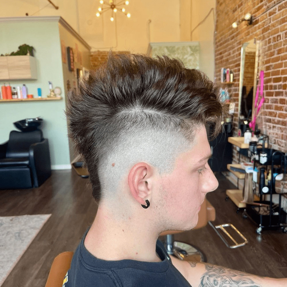 Side profile of a man with a High Fade Mohawk haircut. The hairstyle features a pronounced Mohawk with voluminous, textured hair on top, extending from the forehead to the nape. The sides are shaved into a high fade, creating a stark contrast with the longer hair on top. The man is wearing a black shirt and has an earring, sitting in a modern barbershop with a stylish interior featuring brick walls and contemporary decor. The haircut highlights a bold, edgy look with a clean, sharp fade.