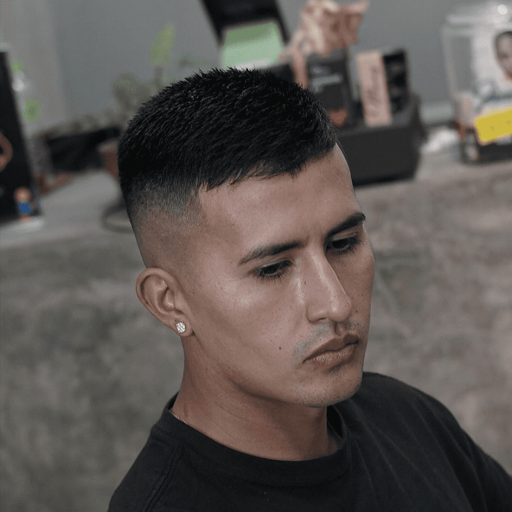 Side profile of a man with a high fade haircut featuring short hair on top. The haircut showcases a clean and sharp fade that starts high on the sides, creating a neat and polished appearance. The man is wearing a black shirt and has a small earring, enhancing his stylish and modern look. The background appears to be a casual indoor setting, adding to the overall relaxed vibe.